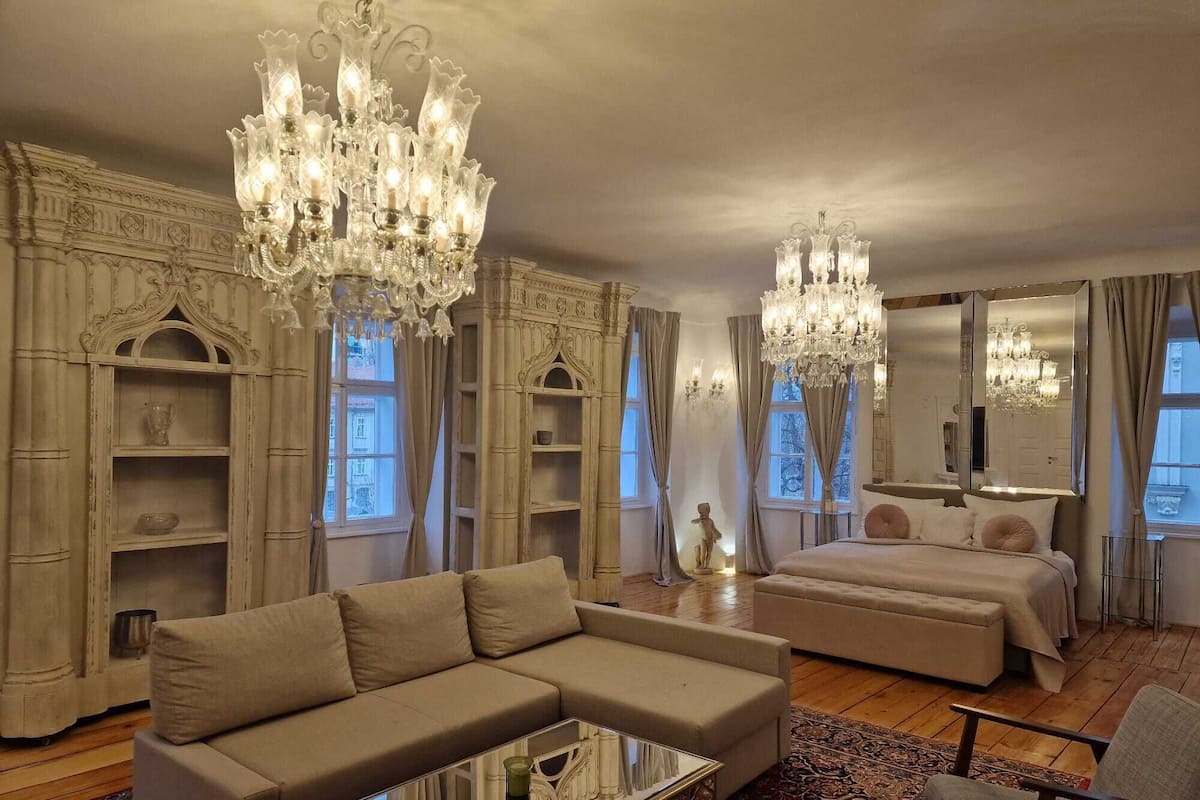 A spacious room with a large bed, a sofa, and a coffee table. There are two chandeliers and a large mirror.