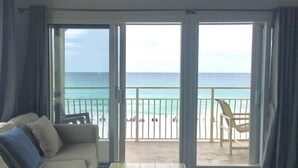 A room with a sofa and a table, a balcony with a view of the ocean and beach, and a chair on the balcony.