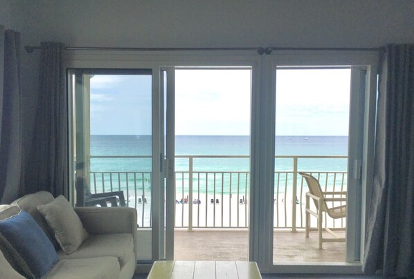 A room with a sofa and a table, a balcony with a view of the ocean and beach, and a chair on the balcony.