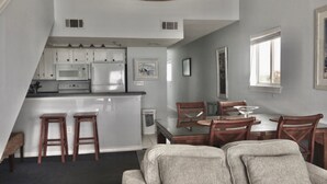 A modern kitchen with white cabinets, a microwave, and a refrigerator. There is a dining area with wooden chairs and a glass table.
