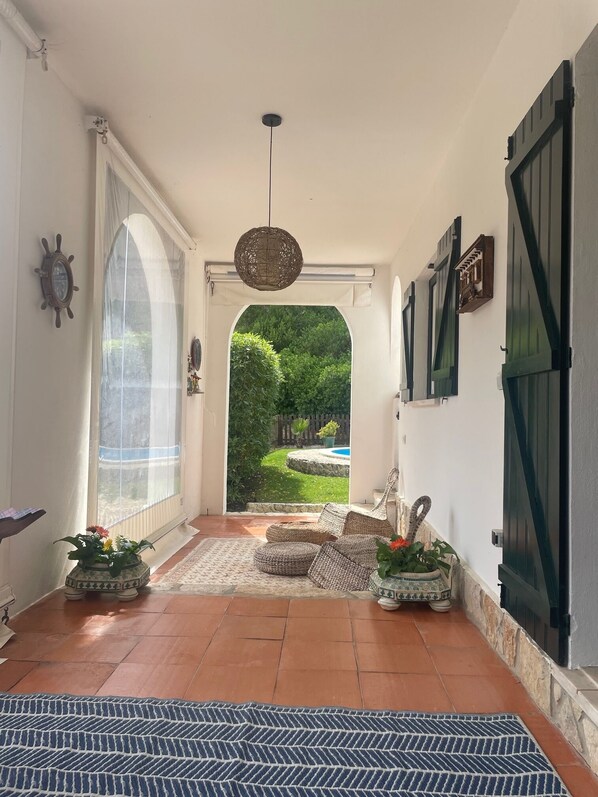 Pasillo con vista a un jardín y una piscina, mobiliario de mimbre y alfombra estampada.