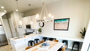 A modern kitchen with white cabinets, a central island, and a dining area with a 'WELCOME' sign. - Ocotillo Springs 49| Private Heated Pool & Spa | 5 Bedroom & 5.5 Bath (SANTA CLARA)