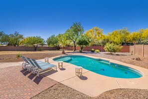 A rectangular swimming pool with a curved edge, surrounded by a brick patio. There are lounge chairs and a small table on the patio.
