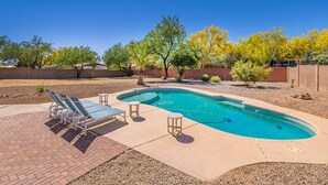 Una piscina rectangular con borde curvo, rodeada por un patio de ladrillo. Hay sillones de descanso y una pequeña mesa cerca de la piscina.