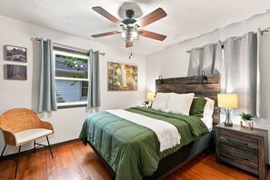 Un dormitorio con cama, una silla, una mesita de noche, un ventilador de techo y una ventana con cortinas.