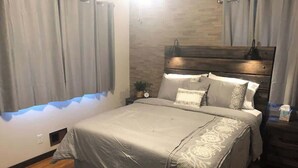 A bedroom with a bed, curtains, a nightstand, a lamp, and a wall-mounted shelf.