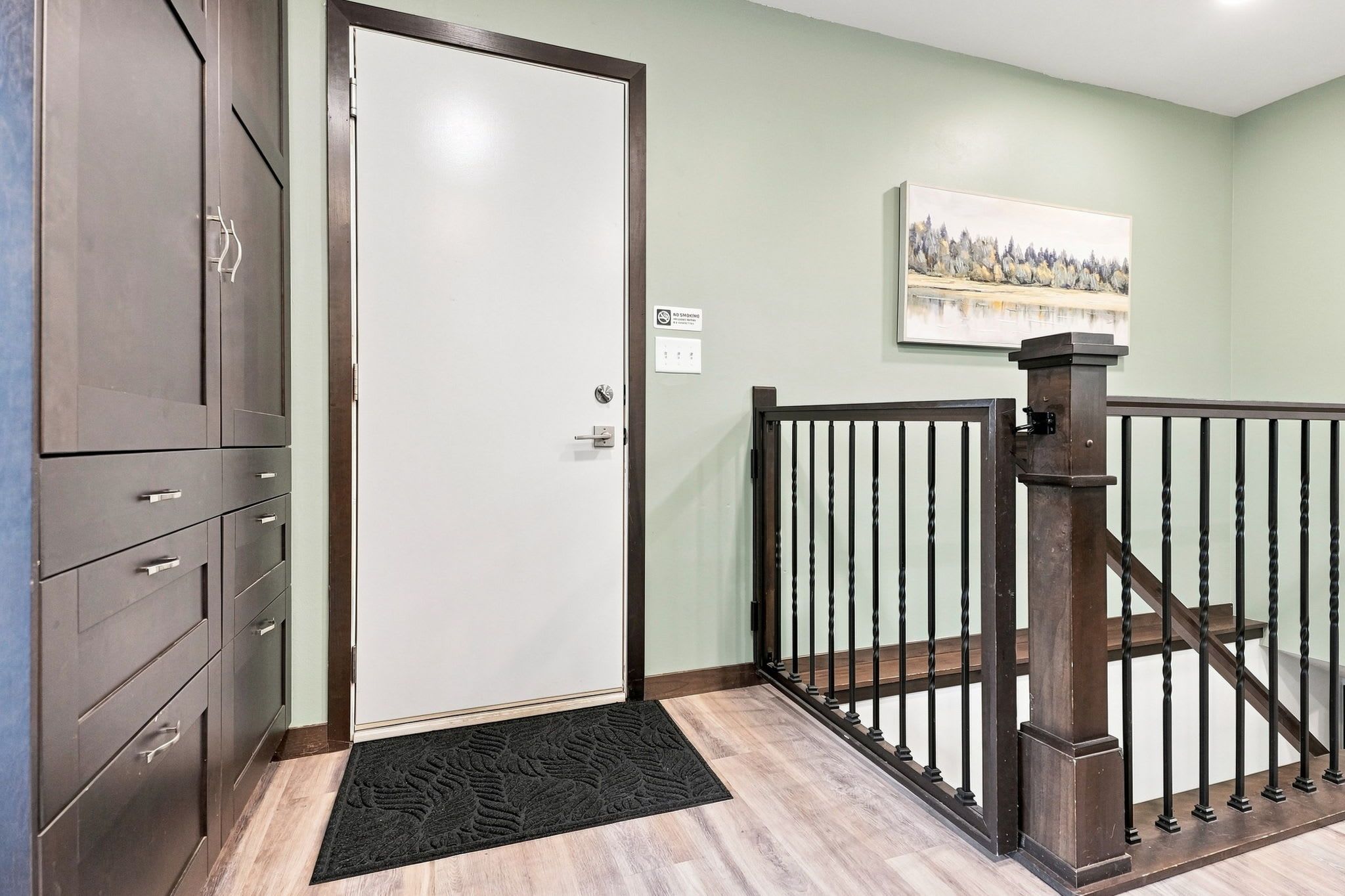Une pièce avec une porte fermée, une commode, un paillasson, un tableau accroché au mur et un escalier avec une rampe.