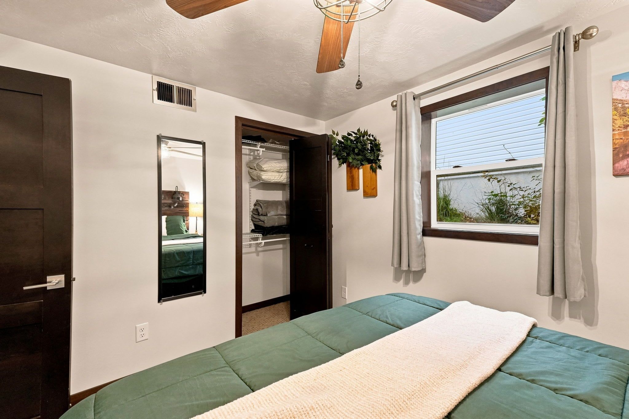 Une chambre à coucher comprenant un lit, une armoire, un miroir, une fenêtre avec des rideaux et un ventilateur de plafond.