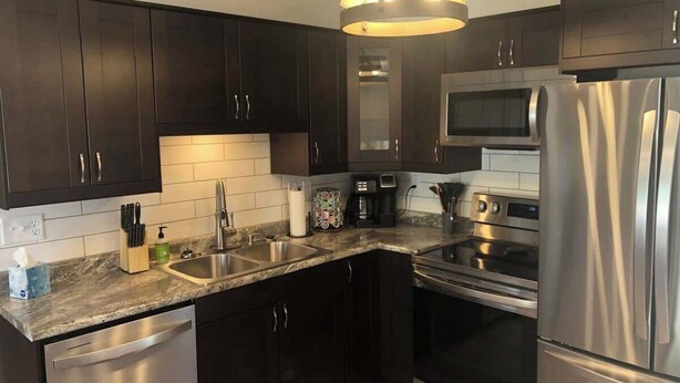 A modern kitchen with dark cabinets, stainless steel appliances, a granite countertop, and a pendant light.