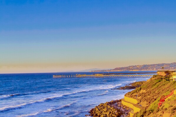 A coastal landscape with a rocky shoreline, a pier extending into the ocean, and a building on a hillside. - New Luxury Condo on Ocean Cliff, 2 Bedroom Suites, Full Kitchen, Living Room (San Diego)