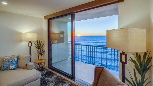 Ein Balkon mit Blick auf das Meer bei Sonnenuntergang, eine Sitzecke, zwei Lampen und ein kleiner Tisch.