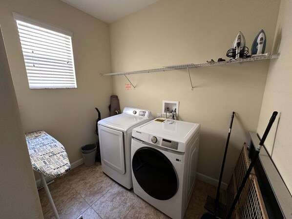 A laundry room with a washing machine, dryer, ironing board, and shelves with ironing boards.