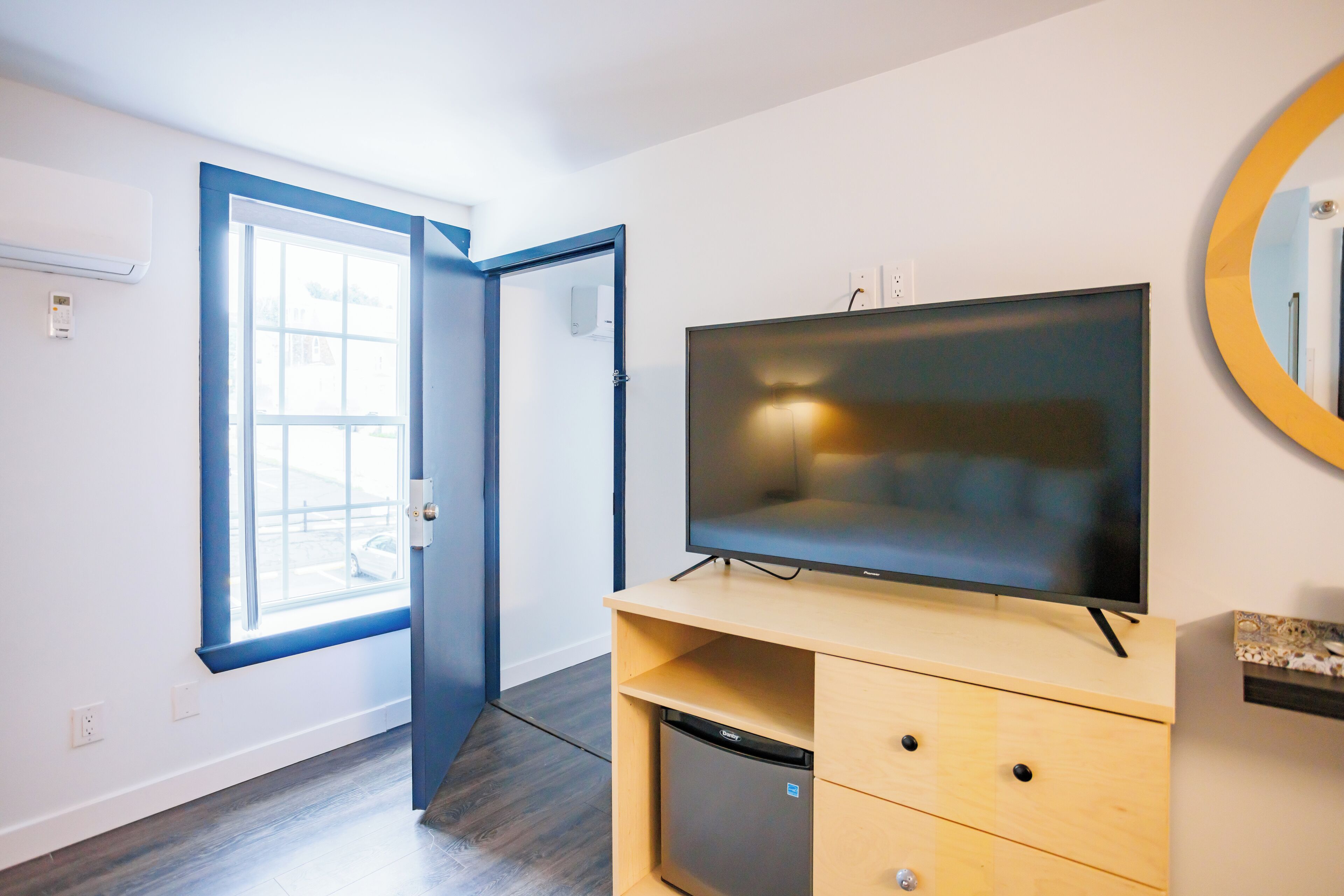 Habitación con un televisor de pantalla plana grande sobre un soporte de madera, un refrigerador pequeño y una puerta que da a un balcón.
