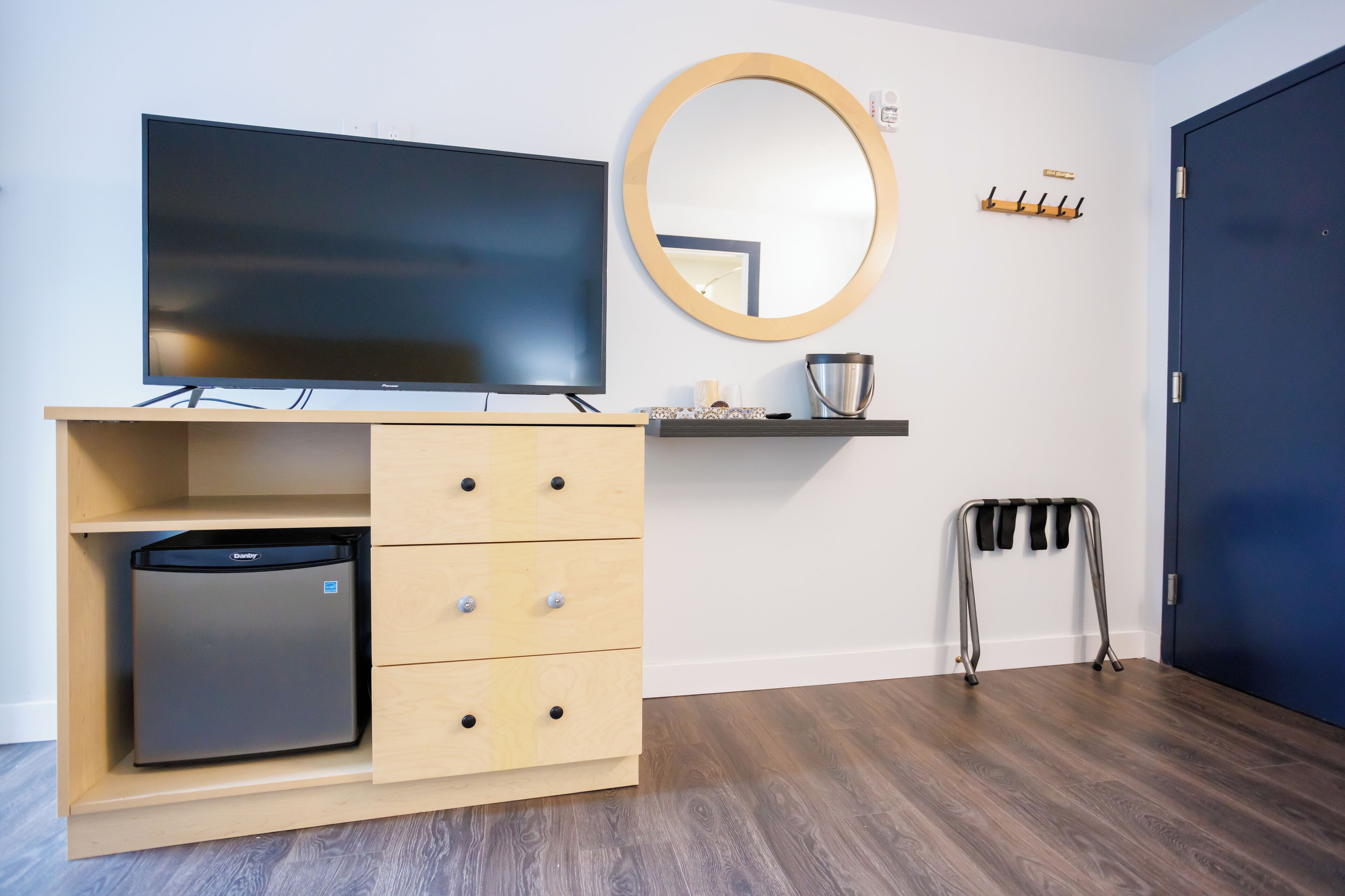 Habitación con televisión de pantalla plana sobre un mueble de madera, un refrigerador pequeño, un espejo y un perchero.