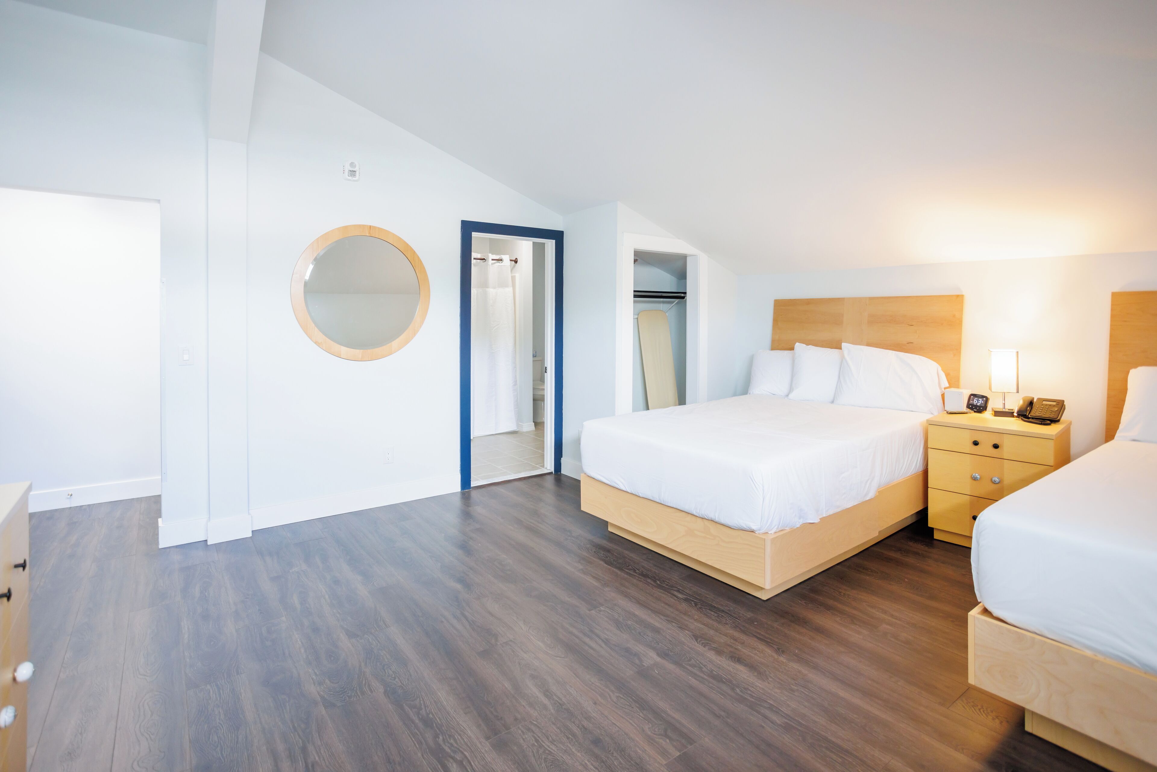 A bedroom with a wooden floor, a bed with white bedding, a bedside table with a lamp, and a mirror on the wall.