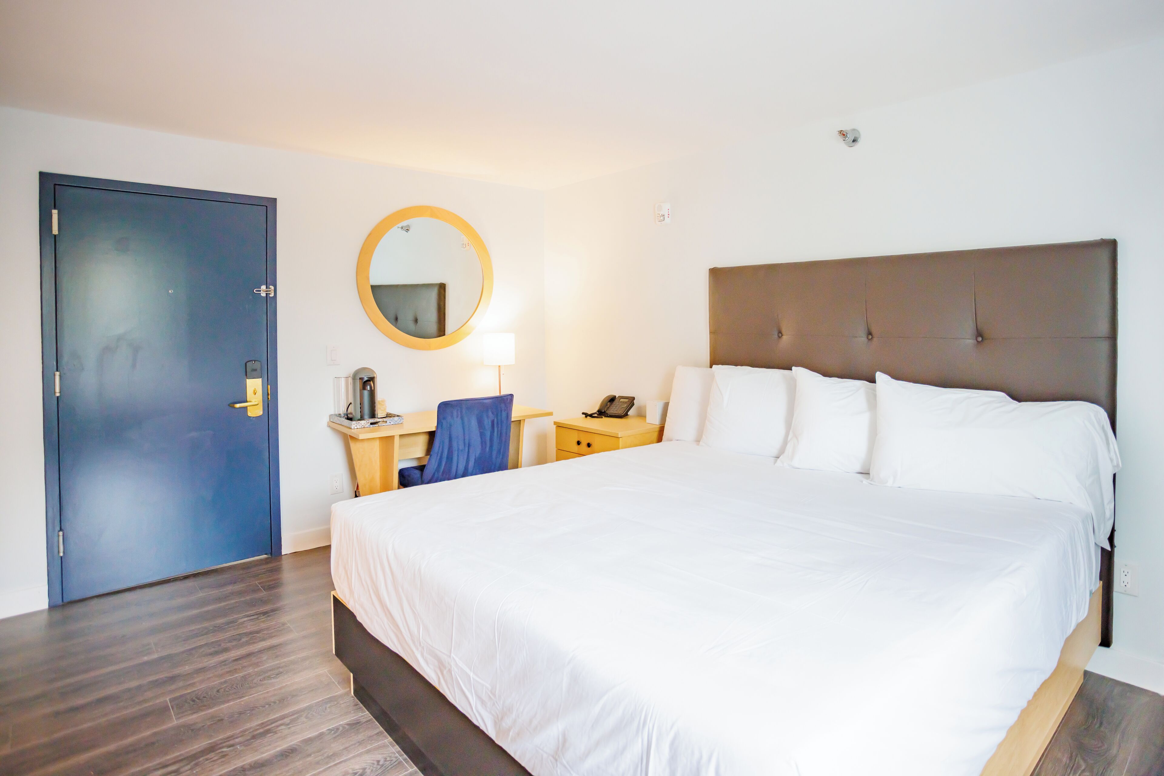 Una habitación de hotel con una cama grande, un escritorio con silla, un espejo redondo y una puerta.