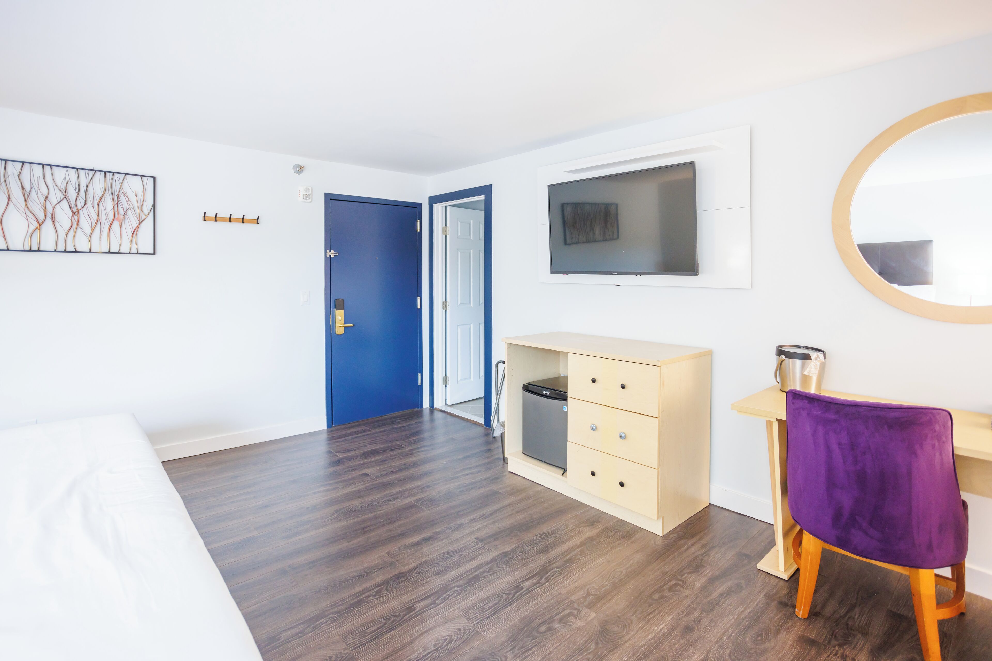 A hotel room with a wooden floor, a bed, a desk with a purple chair, a TV, and a blue door.
