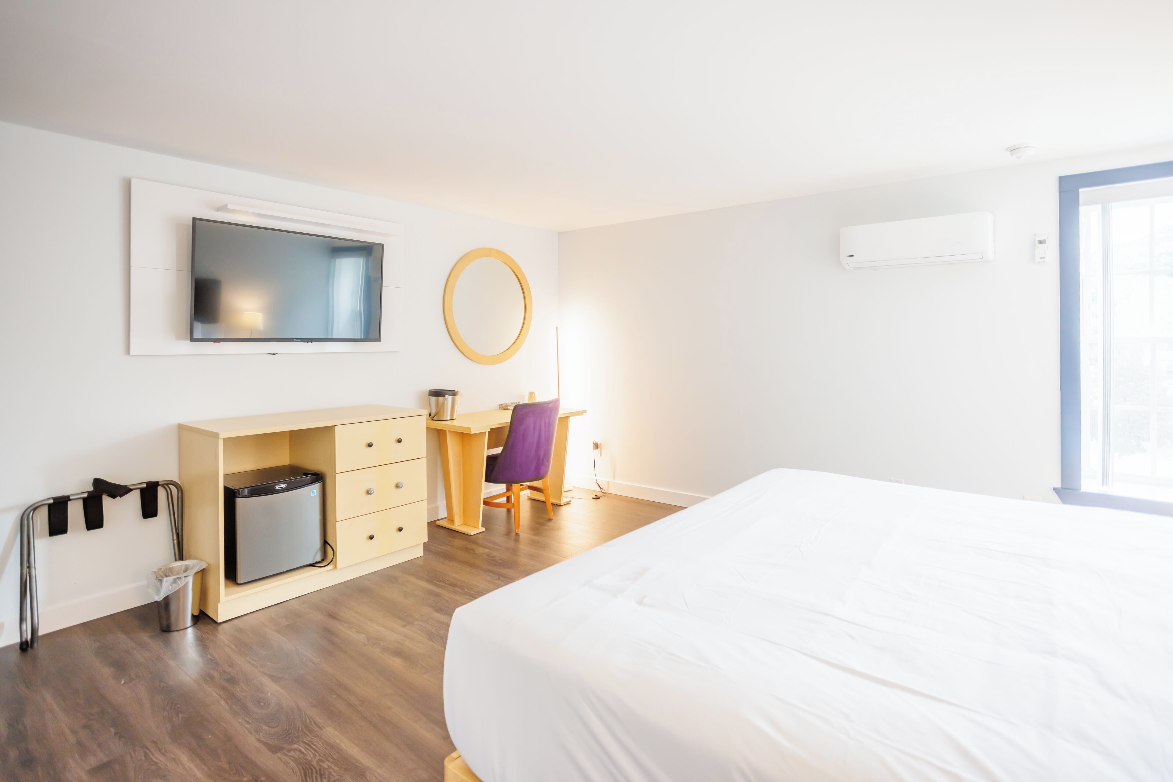 Una habitación de hotel con una cama, un escritorio con una silla, un televisor montado en la pared, un bote de basura pequeño y un equipo de aire acondicionado.