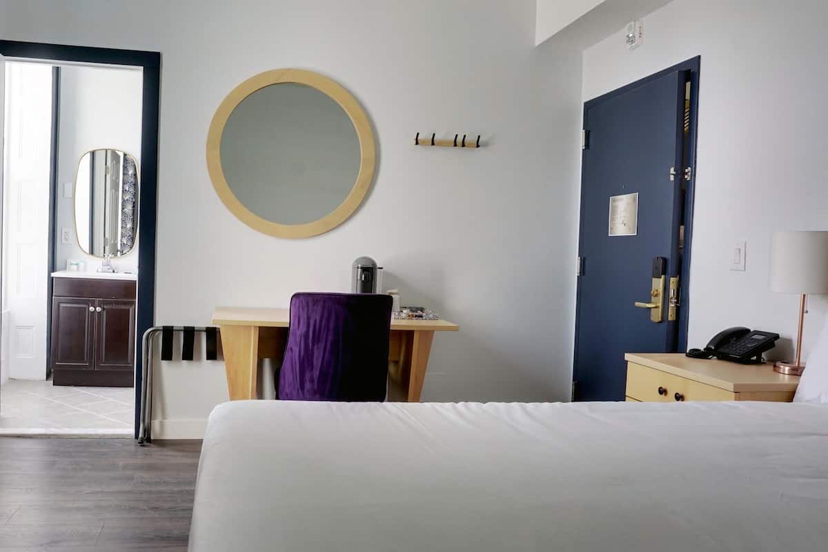 A hotel room with a bed, a desk with a chair, a round mirror, a blue door, and a wooden cabinet.