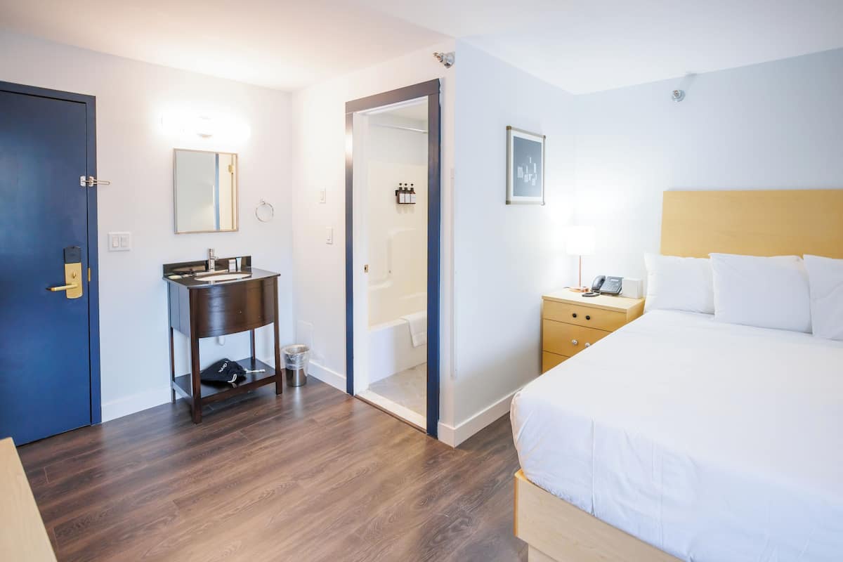 A hotel room with a wooden floor, a bed with white linens, a bedside table with a lamp, a small wooden nightstand, a bathroom with a bathtub, and a door with a blue frame.