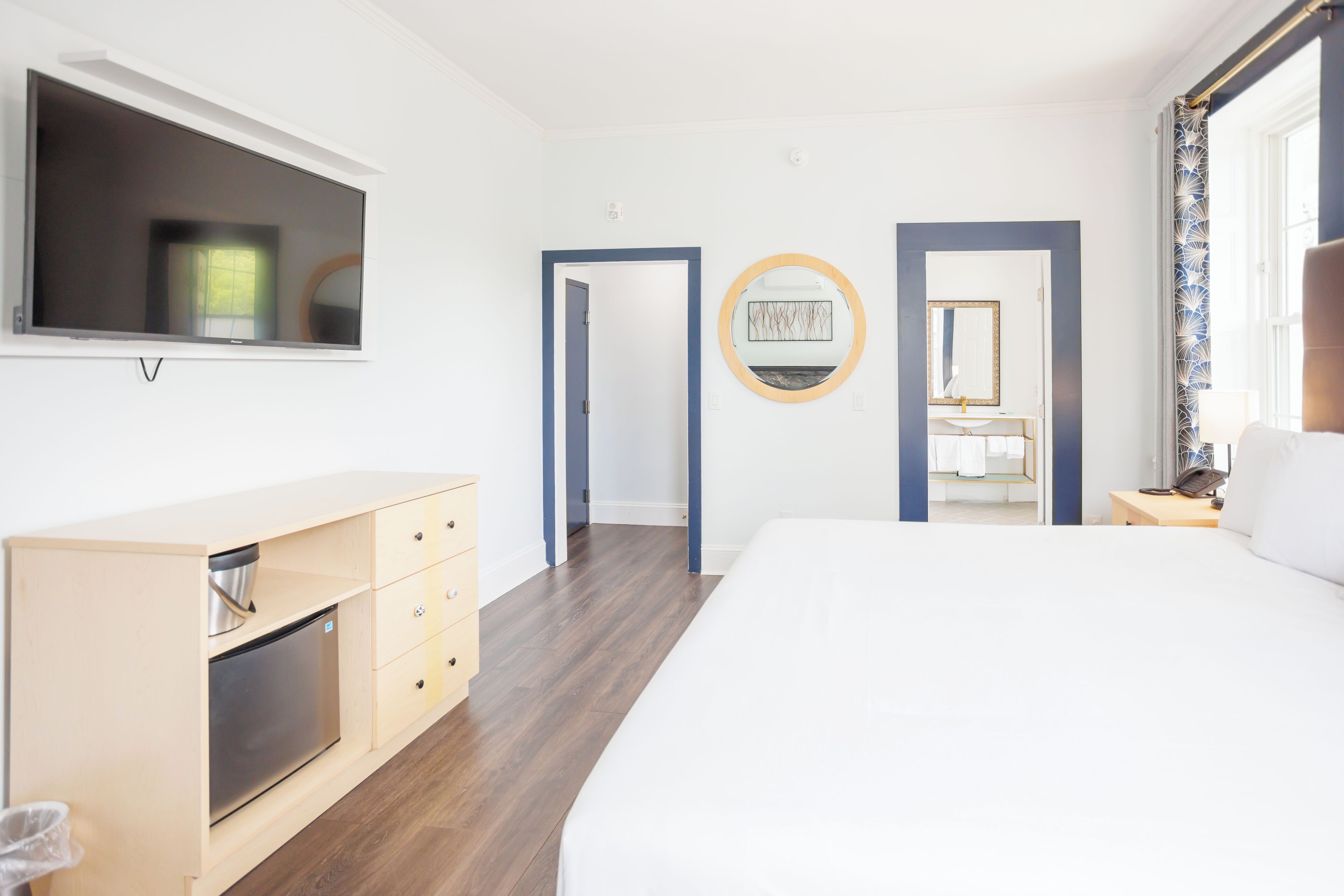 A modern hotel room with a large bed, a TV mounted on the wall, a wooden dresser, and a bathroom visible through an open door.