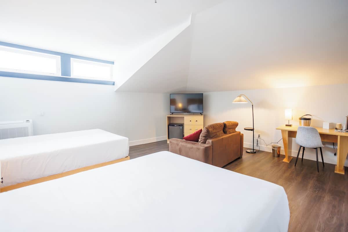 A modern hotel room with two beds, a kitchenette, a desk with a chair, and a large window.