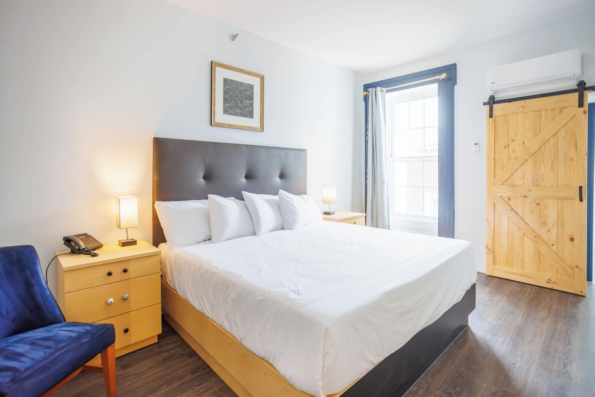 A bedroom with a large bed, bedside tables, a chair, a framed picture on the wall, and a wooden door.