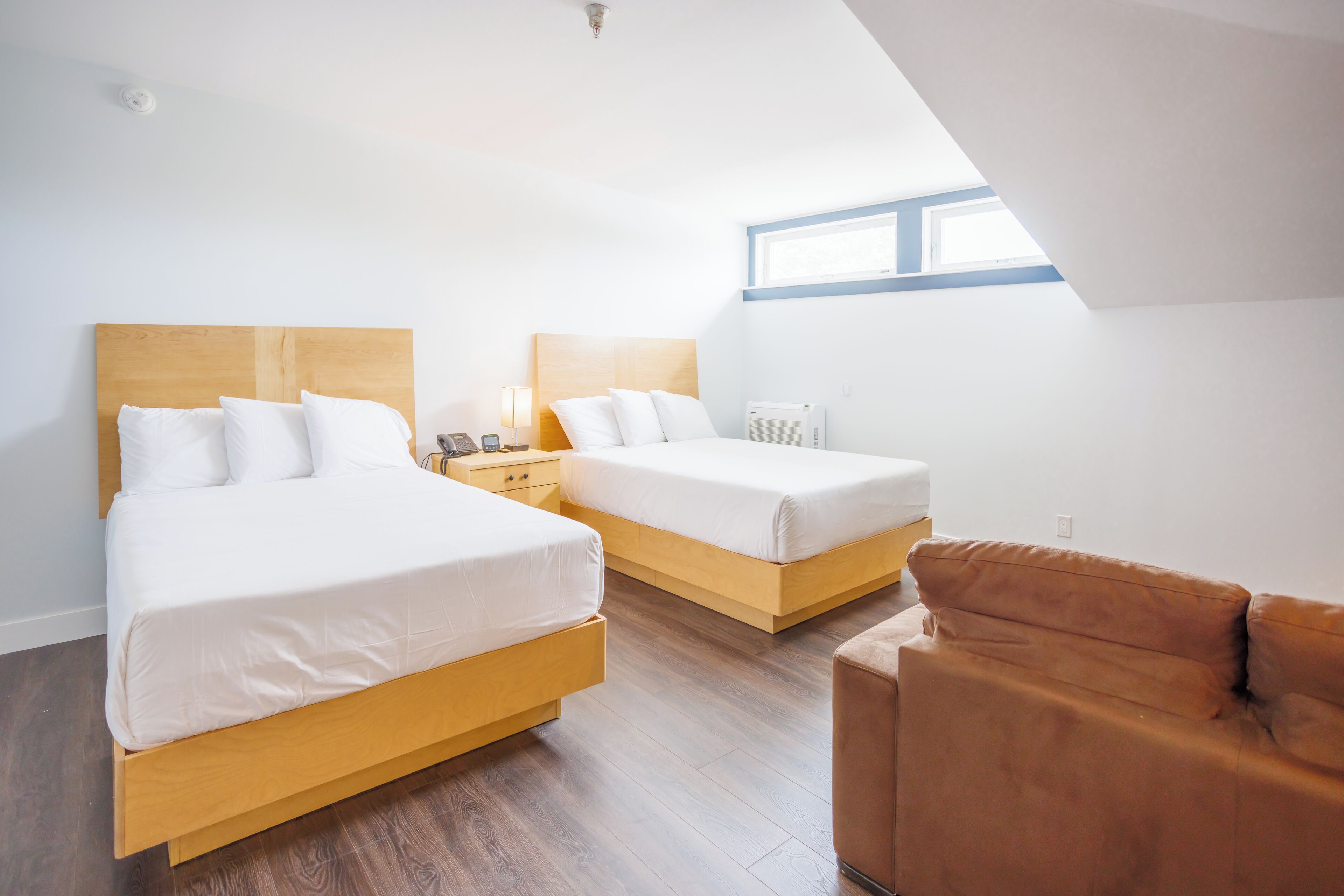 A modern hotel room with two beds, a wooden headboard, a brown leather sofa, and a skylight.