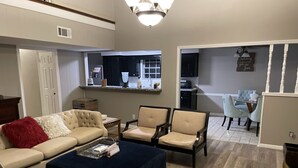 Un salón espacioso con un sofá color beige, dos sillones, una mesa de centro y una zona de comedor con una silla azul y una pequeña mesa.