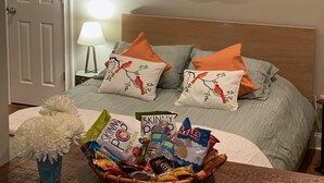 Ein ordentlich bezogenes Bett mit zwei Kissen, ein Nachttisch mit einer Korb voller Snacks und ein Spiegel, der einen Küchenbereich widerspiegelt.