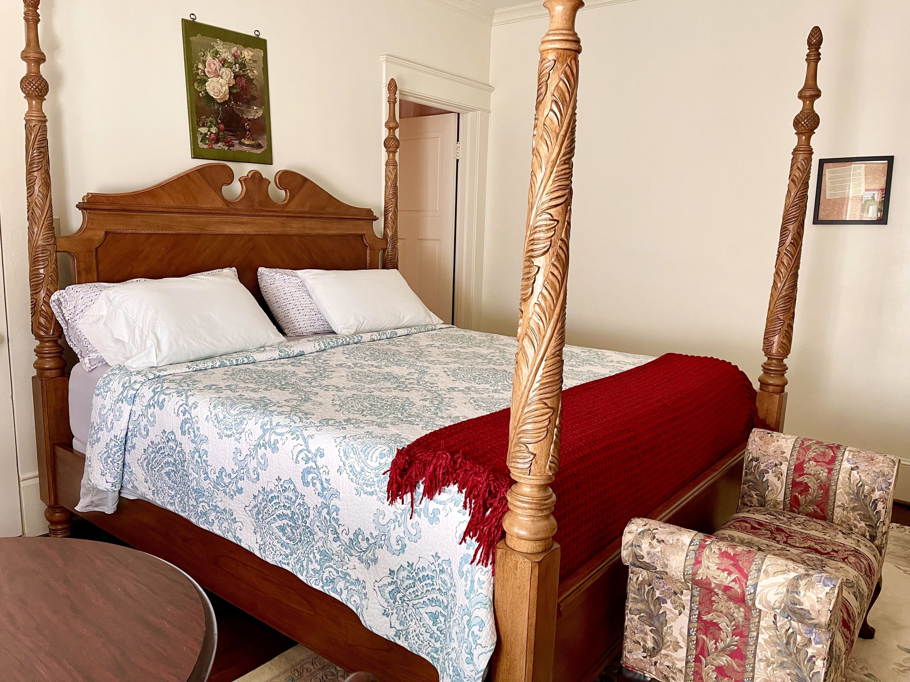 A four-poster bed with a floral bedspread, a red throw blanket, and a matching chair.