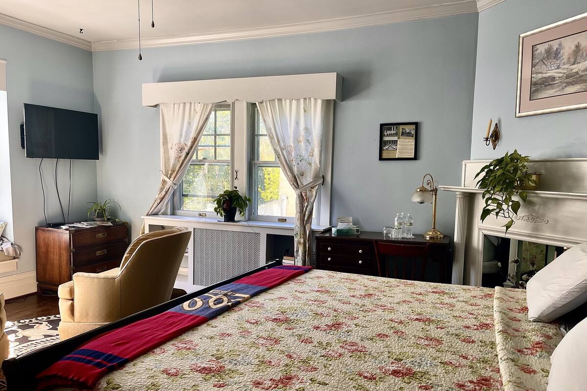 Une chambre à coucher comprenant un lit, un bureau, une chaise, une télévision, une cheminée et une fenêtre avec des rideaux.