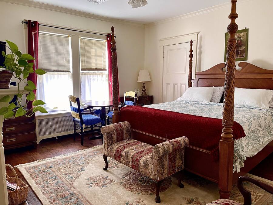 A bedroom with a four-poster bed, a chair, a desk, and a window with curtains.