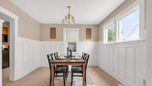 Ein Esszimmer mit Holztisch, gedeckt für vier Personen, weißen Wandverkleidungen, einer Chandelierleuchtre und Blick durch ein Fenster.