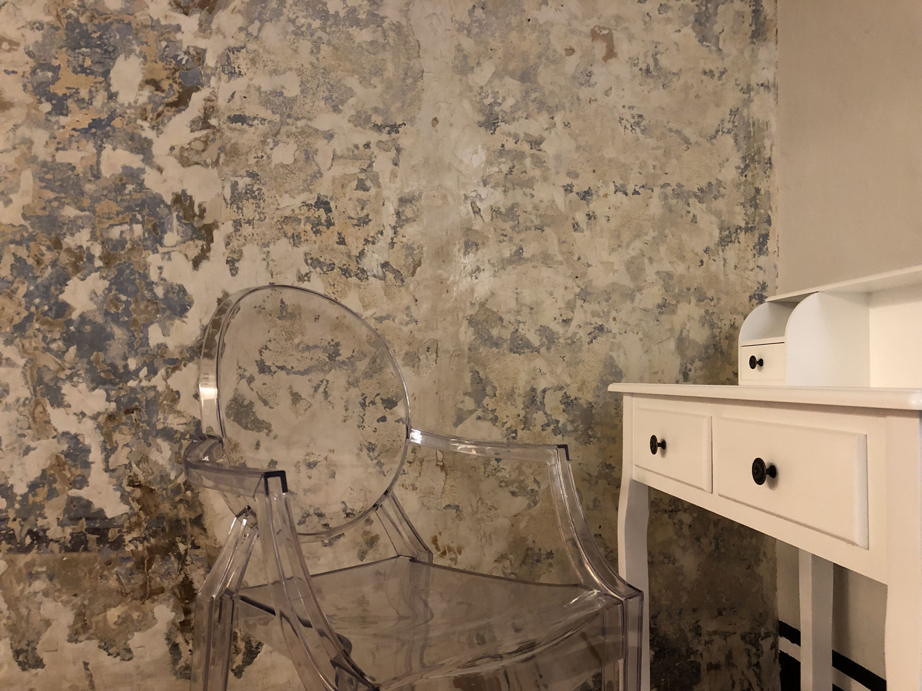 A clear glass chair against a textured wall, next to a white dresser with two drawers.