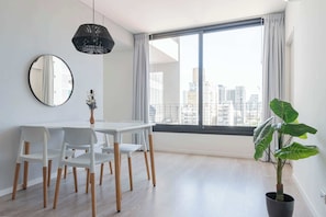 A modern dining area with a white table and chairs, a large window with a city view, a hanging light fixture, and a potted plant.