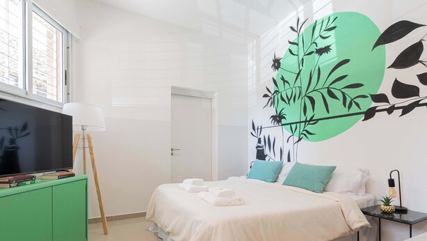 Una habitación de hotel con cama, televisor en un mueble verde, un cuadro de hojas y una ventana.