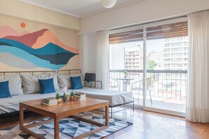 A modern bedroom with a large bed, a sofa, a wooden coffee table, and a view of the city through a sliding glass door.