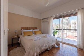 Ein Schlafzimmer mit einem großen Bett, einem Nachttisch, einer Lampe, einem Topf mit Pflanze und Blick auf Gebäude vom Balkon aus.