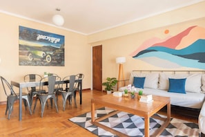 A modern living room with a wooden coffee table, a sofa with blue cushions, a dining area with metal chairs, and a painting of a car and landscape on the wall.