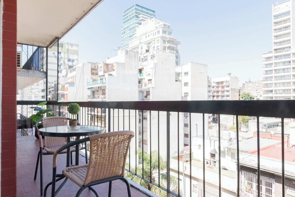 A balcony with wicker chairs and a small table, overlooking a cityscape with modern buildings.