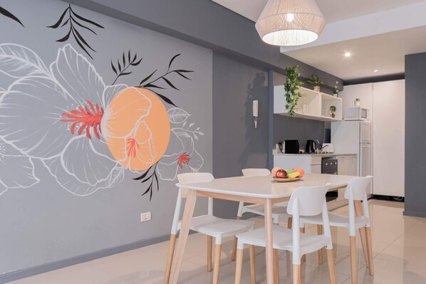 A modern dining area with a white table and chairs, a decorative wall mural, and a kitchen in the background.