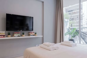 A modern hotel room with a large flat-screen TV mounted on the wall, a neatly made bed with folded towels, a small shelf with books and decorative items, and a potted plant by the window.