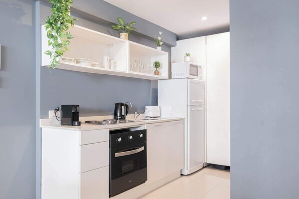 A modern kitchen with white cabinetry, a built-in oven, a microwave, and a coffee maker. There are shelves above the counter holding glassware and a potted plant.