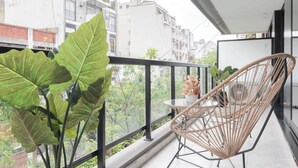 A balcony with a wicker chair and a table, overlooking a cityscape with buildings and greenery.