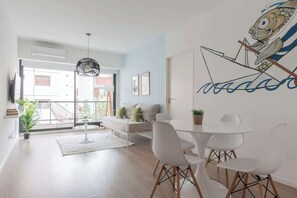 Ein modernes Wohnzimmer mit einem weißen Sofa, einem Esstisch mit weißen Stühlen, einer Pendelleuchte und einer Wandmalerei.