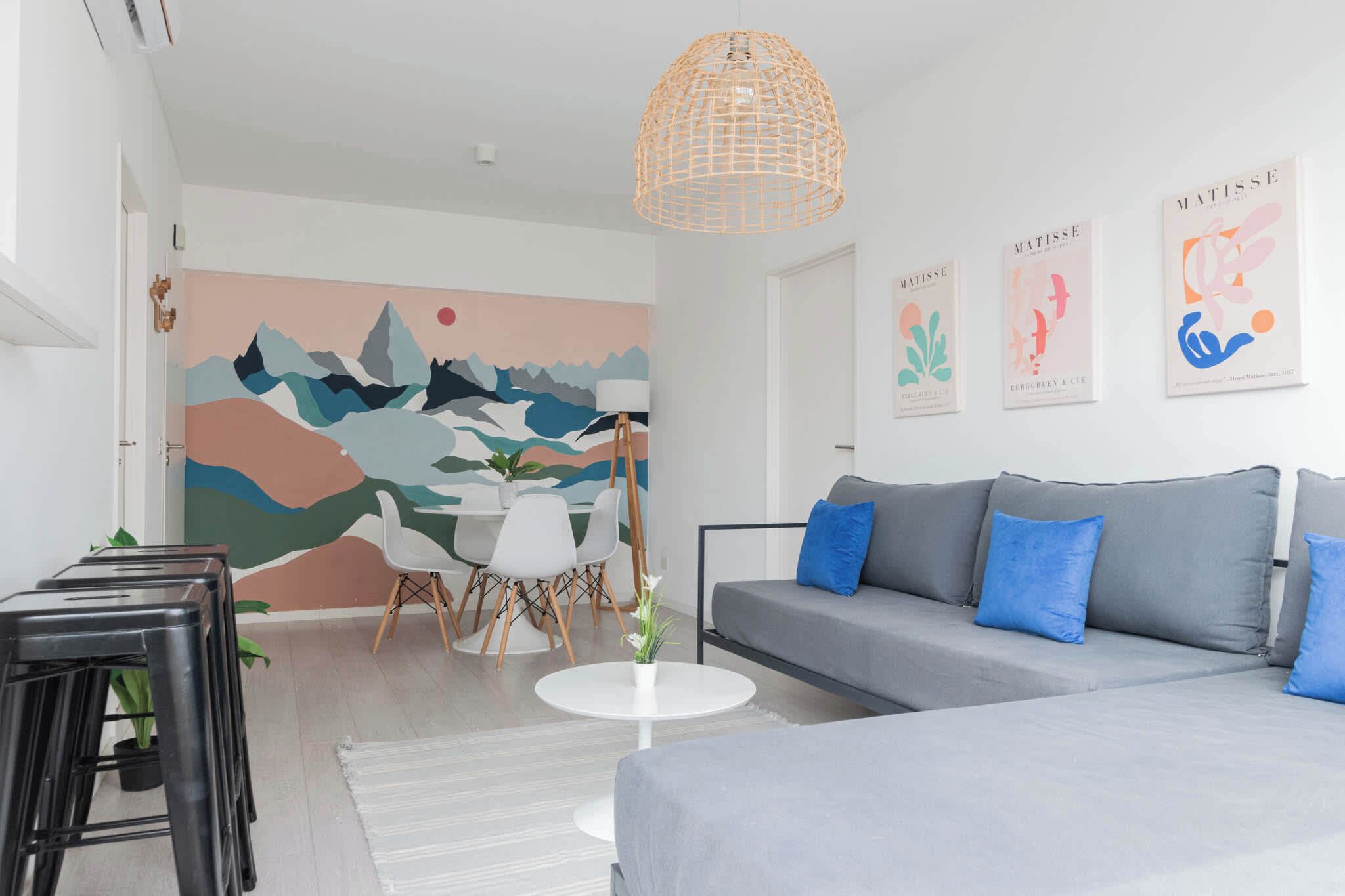 Una sala moderna con un sofá gris, cojines azules, una mesa de centro blanca y un mural abstracto y colorido en la pared.