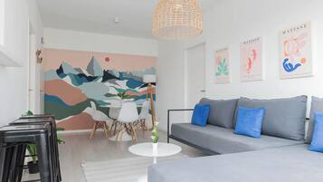 Una sala moderna con un sofá gris, cojines azules, una mesa de centro blanca y un mural abstracto y colorido en la pared.
