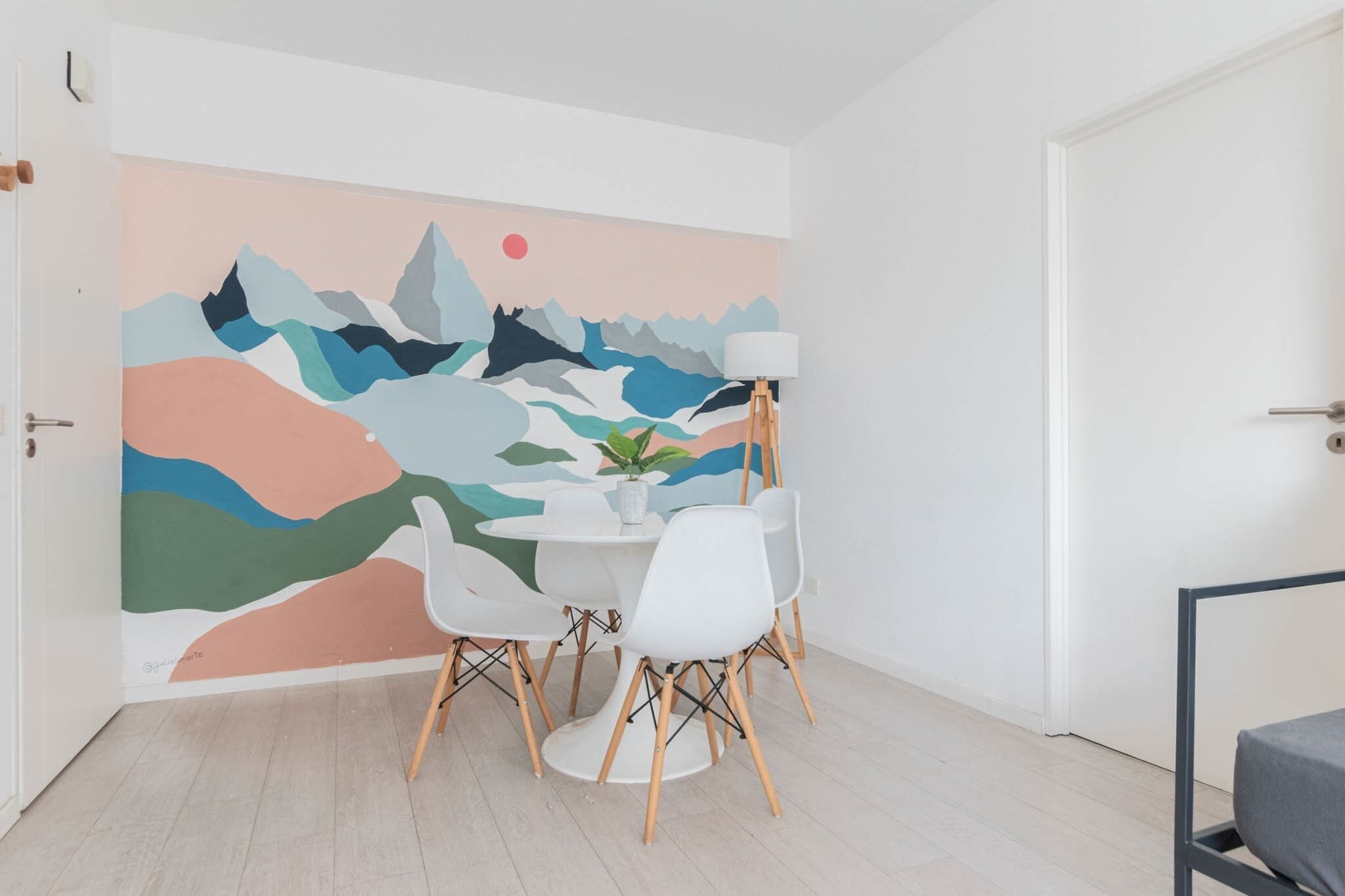 Una habitación con pared blanca, un mural abstracto lleno de color, una mesa redonda con sillas blancas y una lámpara de pie.