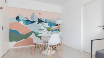 Una habitación con pared blanca, un mural abstracto lleno de color, una mesa redonda con sillas blancas y una lámpara de pie.
